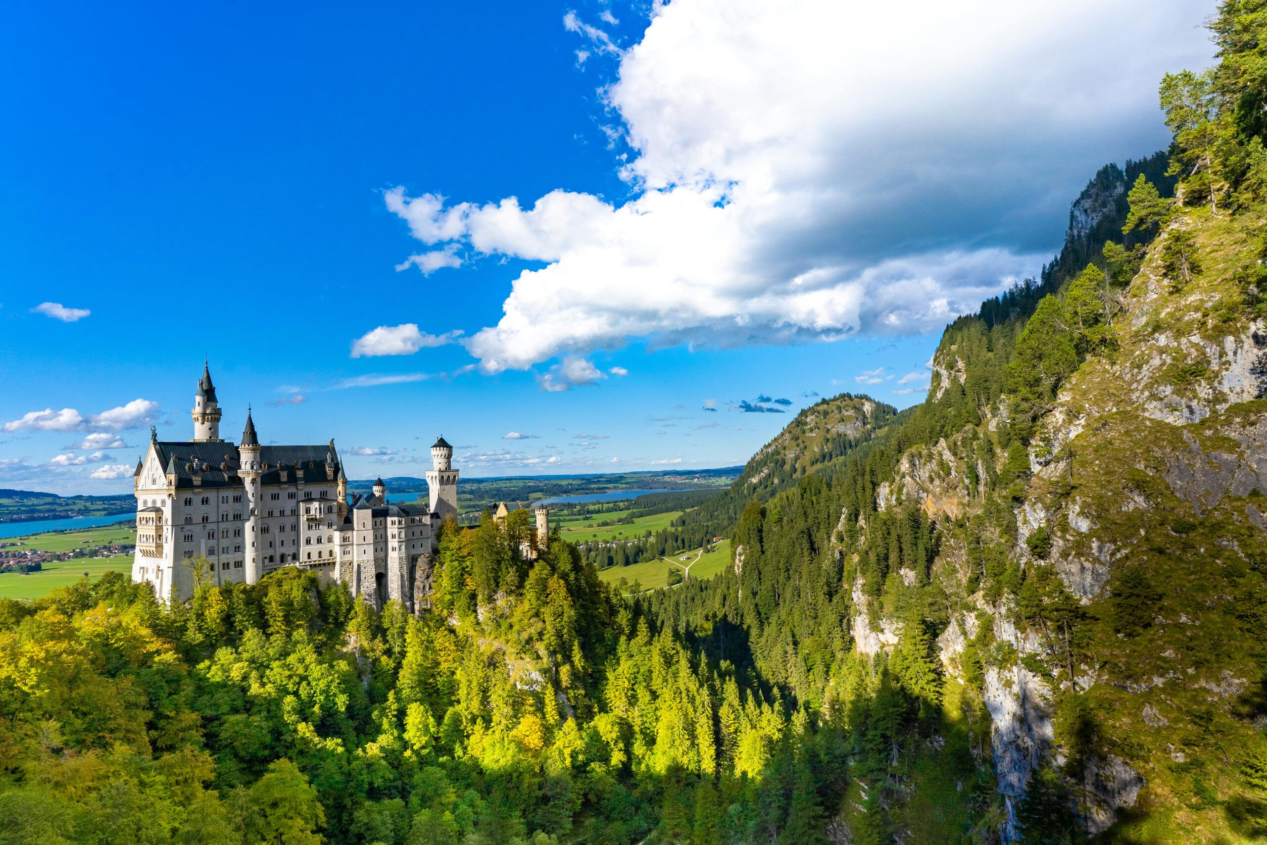 Aerial view of Neuschwanstein Castle in Bavaria, Germany, surrounded by lush forests and mountains under a blue sky.